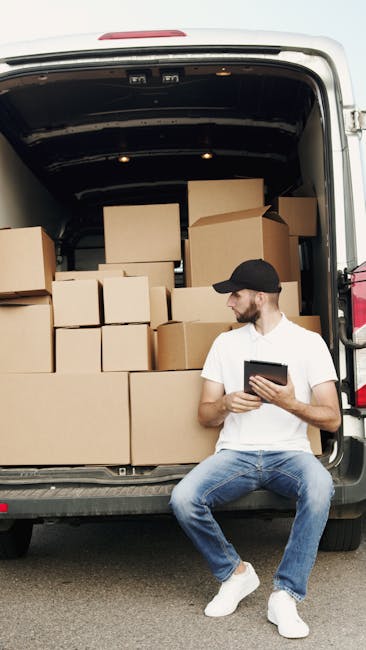 A man wearing a black cap, white T-shirt, and blue jeans is sitting on the pavement beside an open van, which is packed with stacked cardboard boxes of various sizes. The van's interior reveals a load