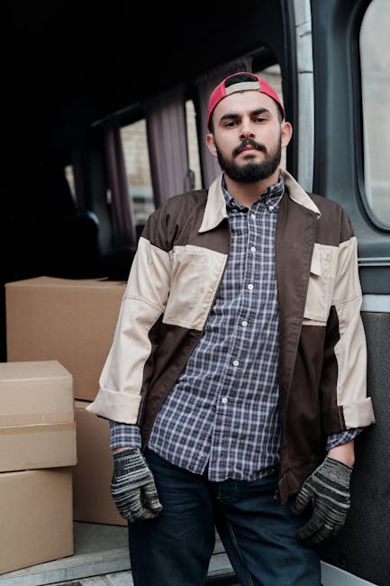 A man wearing a black cap, white T-shirt, and blue jeans is sitting on the pavement beside an open van, which is packed with stacked cardboard boxes of various sizes. The van's interior reveals a load