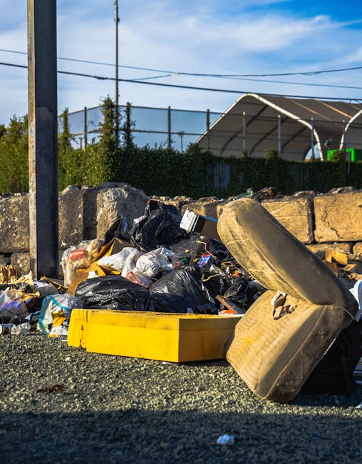 An outdoor scene showing a pile of household waste and discarded items on a gravel surface beside a stone wall, including black plastic garbage bags, a yellow plastic container, and a large, worn sofa cushion leaning against the rubbish. Behind the debris, a metal pole and overhead cables are visible, along with a blue sky and some trees and a roofed structure in the background. The setting suggests a waste disposal area or skip site, with no furniture being actively loaded or transported in the current moment, which is part of the bulky waste removal process managed by Man With a Van Woolwich as part of their house removals and relocation services.