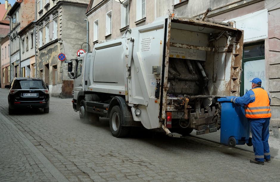 A street scene showing a white rubbish collection truck with an open rear compartment positioned adjacent to a curb in Woolwich. A waste collector, dressed in a high-visibility orange vest, blue uniform, and a blue cap, is emptying a blue wheelie bin into the truck's hopper, which contains various materials including cardboard and plastic. The truck is parked on a cobblestone road alongside residential buildings with multi-paned windows and some brickwork visible. Nearby, a black car is parked facing away from the camera, and the area appears to be set during daytime with natural lighting. This image depicts a typical home waste collection process, which is part of the broader house removal and relocation services offered by Man With a Van Woolwich, supporting efficient packing and transport logistics.