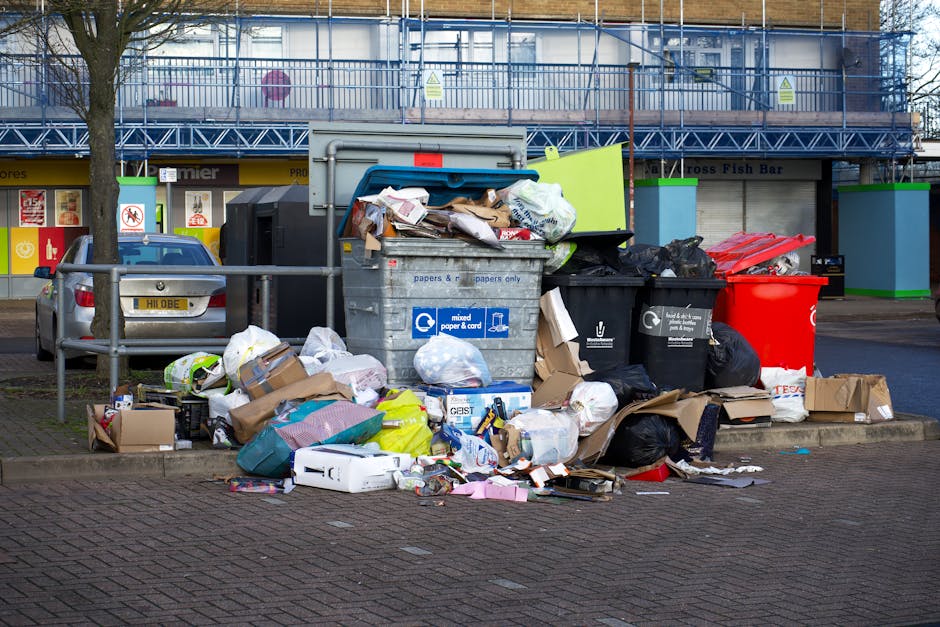 An outdoor scene showing a pile of household waste and discarded items on a gravel surface beside a stone wall, including black plastic garbage bags, a yellow plastic container, and a large, worn sofa cushion leaning against the rubbish. Behind the debris, a metal pole and overhead cables are visible, along with a blue sky and some trees and a roofed structure in the background. The setting suggests a waste disposal area or skip site, with no furniture being actively loaded or transported in the current moment, which is part of the bulky waste removal process managed by Man With a Van Woolwich as part of their house removals and relocation services.