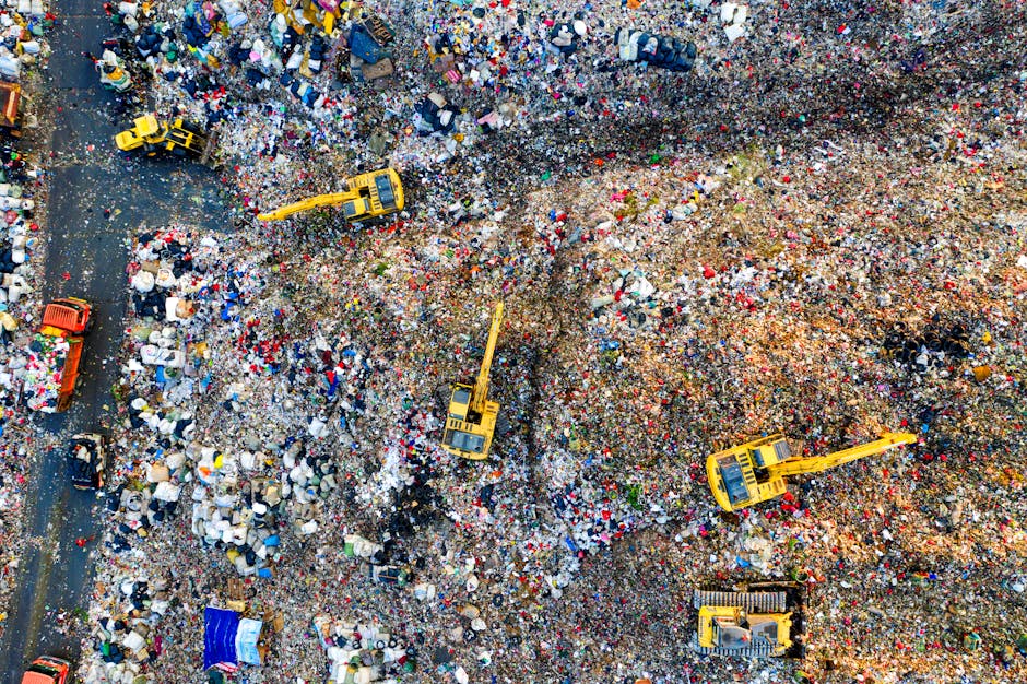 An aerial view of a landfill site filled with scattered household waste, including plastic bottles, cardboard boxes, fabric, and other rubbish. Several yellow excavators with long arms are actively engaged in the process of sorting and moving debris, positioned across the site. A small section of a paved road is visible on the left side, partially covered by waste, with a few vehicles present. The scene depicts a busy waste management operation, with large piles of mixed refuse and machinery working amidst the cluttered environment. This image emphasizes the scale of waste disposal and the importance of proper rubbish removal in household relocation and moving services, such as those offered by Man With a Van Woolwich, to support efficient house removals and packing preparations.