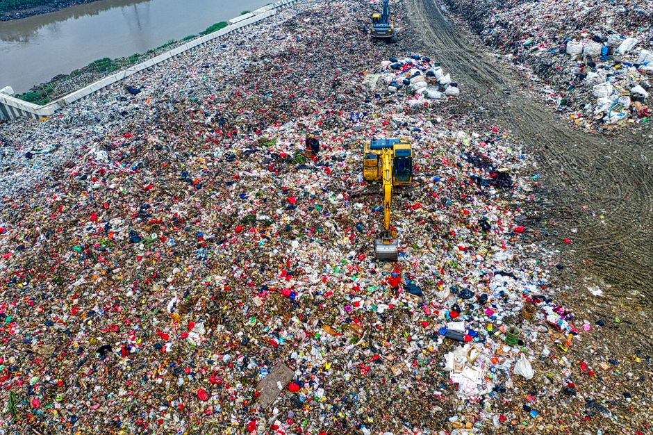 An aerial view of a large landfill site filled with mixed household and commercial waste, including plastic bottles, cardboard boxes, and plastic packaging. A yellow excavator is actively moving and sorting through the piled debris on a dirt-covered area. The site is surrounded by a gravel pathway and a barrier wall, with the landscape showing signs of extensive waste accumulation. The image captures a scene of waste disposal and rubbish management, relevant to home relocation waste handling and disposal rules, as discussed on the page titled 'Council Waste Rules in Woolwich' by Man With a Van Woolwich, a company offering removals and moving services.