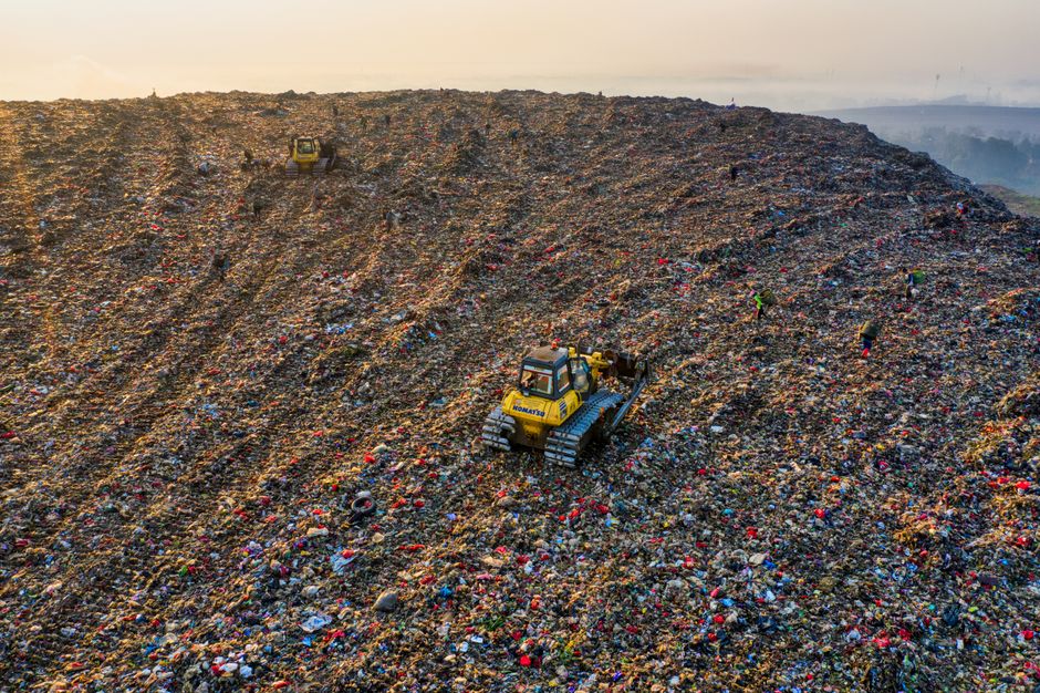 An aerial view of a landfill site filled with scattered household waste, including plastic bottles, cardboard boxes, fabric, and other rubbish. Several yellow excavators with long arms are actively engaged in the process of sorting and moving debris, positioned across the site. A small section of a paved road is visible on the left side, partially covered by waste, with a few vehicles present. The scene depicts a busy waste management operation, with large piles of mixed refuse and machinery working amidst the cluttered environment. This image emphasizes the scale of waste disposal and the importance of proper rubbish removal in household relocation and moving services, such as those offered by Man With a Van Woolwich, to support efficient house removals and packing preparations.