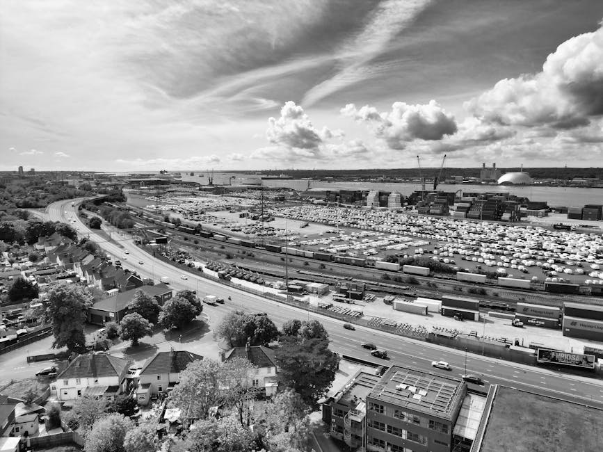 A high-angle, black-and-white aerial photograph showing a large industrial area with numerous shipping containers, warehouses, and cranes, indicating active freight handling and logistics operations near a body of water in Woolwich. In the foreground, residential houses with pitched roofs and surrounding trees are visible, along with a multi-lane road carrying moving traffic. The scene captures the contrast between urban residences and commercial transport infrastructure, illustrating the scale of logistics involved in home relocation and furniture transport services provided by Man With a Van Woolwich, with the focus on the exterior environment, loading and unloading zones, and transport vehicles associated with removals.
