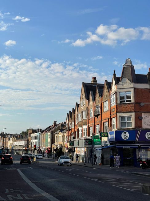 A wide street scene in Woolwich showing a row of residential buildings with brick exteriors and multiple bay windows along the upper floors, under a partly cloudy sky. On the street level, there are commercial shops with signage, including a pharmacy and various other storefronts, alongside pedestrians walking on the sidewalk. Vehicles, including cars and a bus, are moving along the road, while some cars are waiting at a traffic light. The scene captures a typical urban environment where home relocation and furniture transport services, such as those offered by Man With a Van Woolwich, may operate, with the loading process likely happening nearby or in preparation within the vicinity.