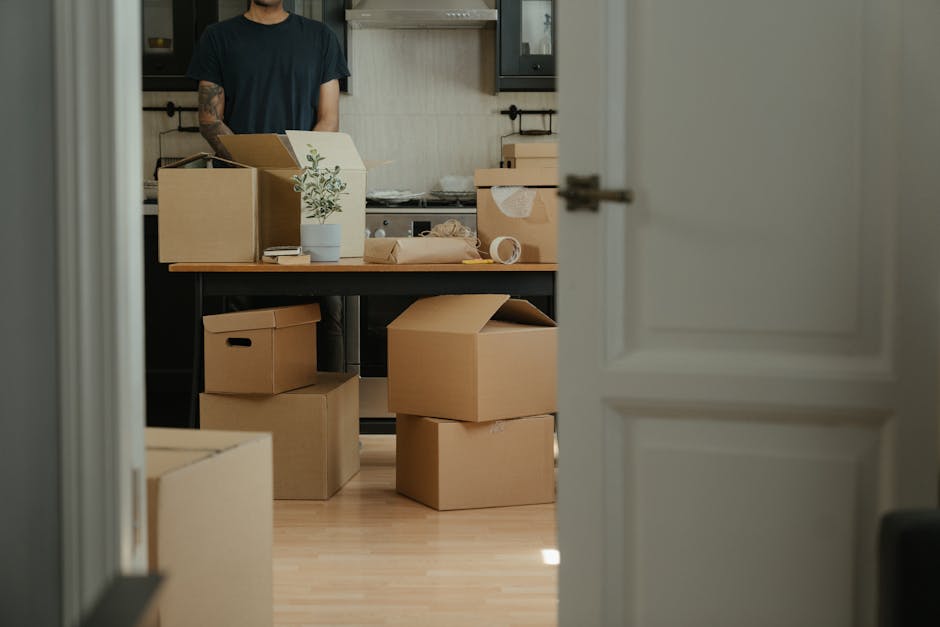 The image shows a domestic kitchen interior viewed through a partially open door, with the focus on a cluttered table filled with packed cardboard boxes, some sealed with packing tape, and a small potted plant. In the background, a person dressed in a black t-shirt is partially visible, standing behind the table, possibly preparing for a house removal or relocation. The kitchen features dark cabinetry, a light-colored wall, and a wooden work surface. Several packed boxes are placed on the light-colored wooden floor, indicating an ongoing packing or moving process. The scene depicts the early stages of a home relocation, with packing materials and boxes prepared for transport. This setup aligns with services offered by Man With a Van Woolwich, focusing on packing, furniture transport, and loading activities involved in efficient house removals within Woolwich.