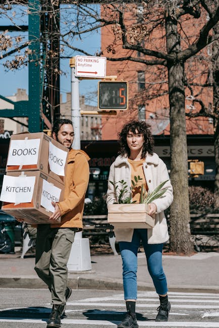 Two individuals walking across a city street during daylight, carrying packed boxes labeled 'BOOKS' and 'KITCHEN'. The man on the left, with dark hair and a beard, wears a mustard jacket and grey trousers, holding a large cardboard box with both hands. The woman on the right, with curly dark hair, wears a white coat over a mustard top and blue jeans, holding a wooden crate with green foliage inside. In the background, there are trees with bare branches, a red-brick building, a traffic light displaying a red hand and a countdown timer showing 5 seconds, a street sign indicating no parking anytime, and part of a parked vehicle. This scene illustrates a home relocation process as part of a house removals service offered by Man With a Van Woolwich, highlighting the logistics of packing and moving during furniture transport.