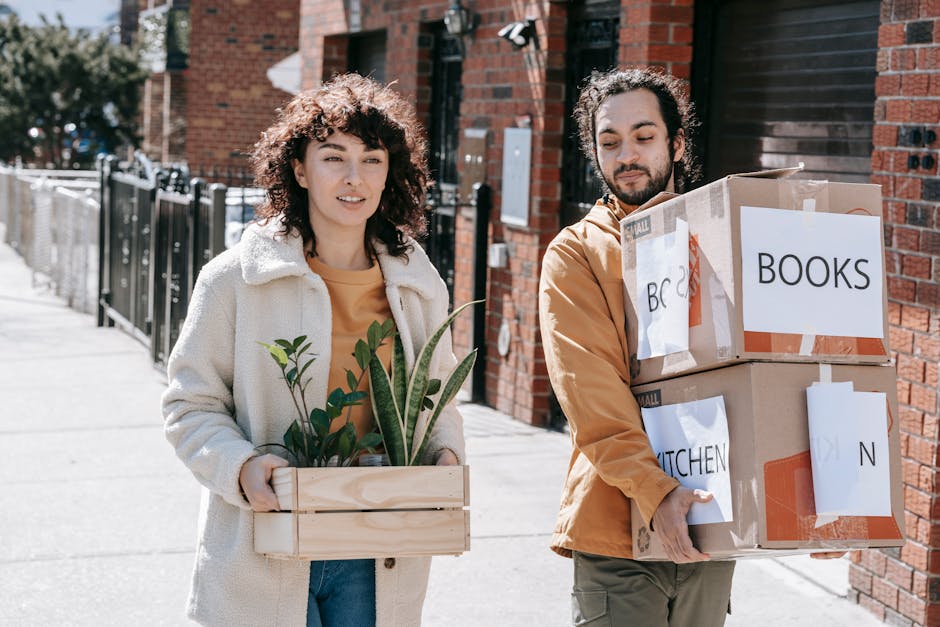 Two individuals walking across a city street during daylight, carrying packed boxes labeled 'BOOKS' and 'KITCHEN'. The man on the left, with dark hair and a beard, wears a mustard jacket and grey trousers, holding a large cardboard box with both hands. The woman on the right, with curly dark hair, wears a white coat over a mustard top and blue jeans, holding a wooden crate with green foliage inside. In the background, there are trees with bare branches, a red-brick building, a traffic light displaying a red hand and a countdown timer showing 5 seconds, a street sign indicating no parking anytime, and part of a parked vehicle. This scene illustrates a home relocation process as part of a house removals service offered by Man With a Van Woolwich, highlighting the logistics of packing and moving during furniture transport.