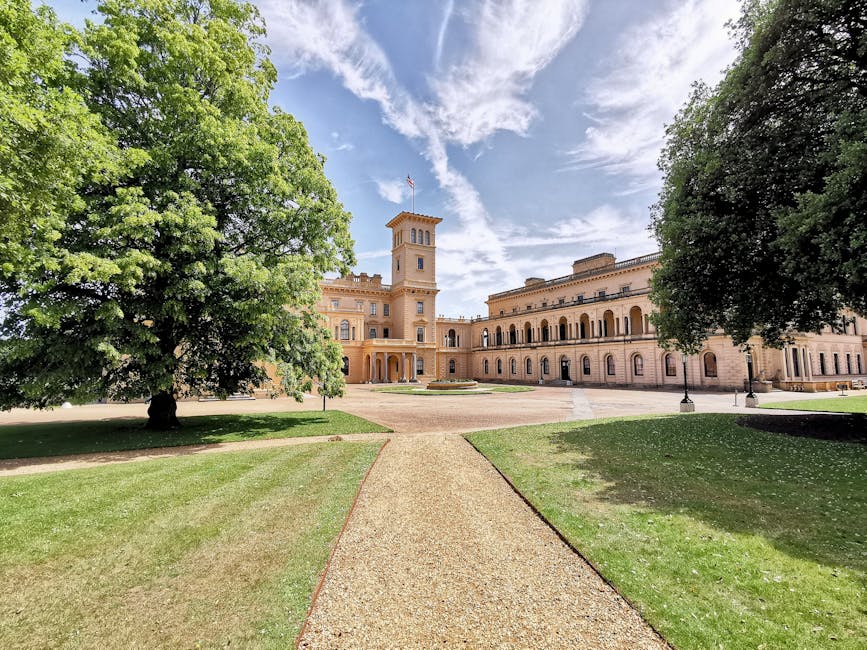 A wide view of a historic sandstone building featuring a central clock tower, set within a landscaped area with a gravel pathway leading directly to the main entrance. On either side of the pathway, t