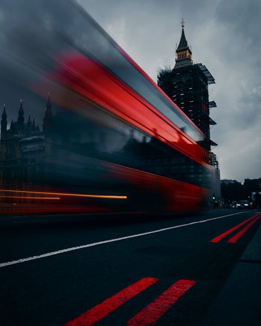 A blurred red and black double-decker bus moving swiftly along a road in London, with the historic Big Ben clock tower and the Houses of Parliament visible in the background. The bus appears to be in motion, creating streaks of light and a sense of speed, while the surrounding environment includes dark pavement with red road markings, suggesting an urban setting during dusk or early evening. The image captures the dynamic nature of city transport and may relate to moving logistics or transportation services offered by Man With a Van Woolwich, highlighting the importance of quick and efficient home relocation or furniture transport in the capital.