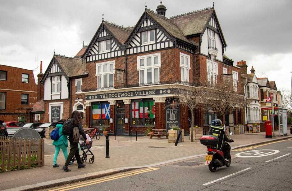 A view of a multi-story brick building with a traditional black and white timber-frame facade, serving as a pub called The Rookwood Village, located on a busy street in Woolwich. The building has multiple windows, some with decorative ironwork balconies, and flags hanging from the front. In front of the pub, a woman with long dark hair, wearing a black jacket and turquoise pants, is walking with a person pushing a stroller. To the right, a delivery rider wearing a helmet and high-visibility vest is sitting on a black scooter parked on the street, near a red public telephone booth and a red post box. The street scene includes bicycles parked nearby, a small fenced garden area with a bench and a leafless tree, and a bus stop with a shelter on the sidewalk, all under a cloudy sky. The setting depicts the busy environment around a local pub, relevant to house removals and moving logistics, illustrating parking and pedestrian activity in Woolwich.