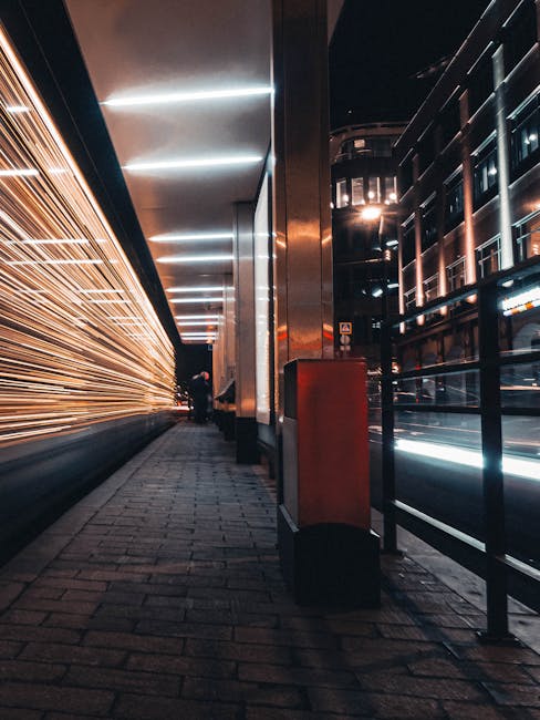 A blurred red and black double-decker bus moving swiftly along a road in London, with the historic Big Ben clock tower and the Houses of Parliament visible in the background. The bus appears to be in motion, creating streaks of light and a sense of speed, while the surrounding environment includes dark pavement with red road markings, suggesting an urban setting during dusk or early evening. The image captures the dynamic nature of city transport and may relate to moving logistics or transportation services offered by Man With a Van Woolwich, highlighting the importance of quick and efficient home relocation or furniture transport in the capital.