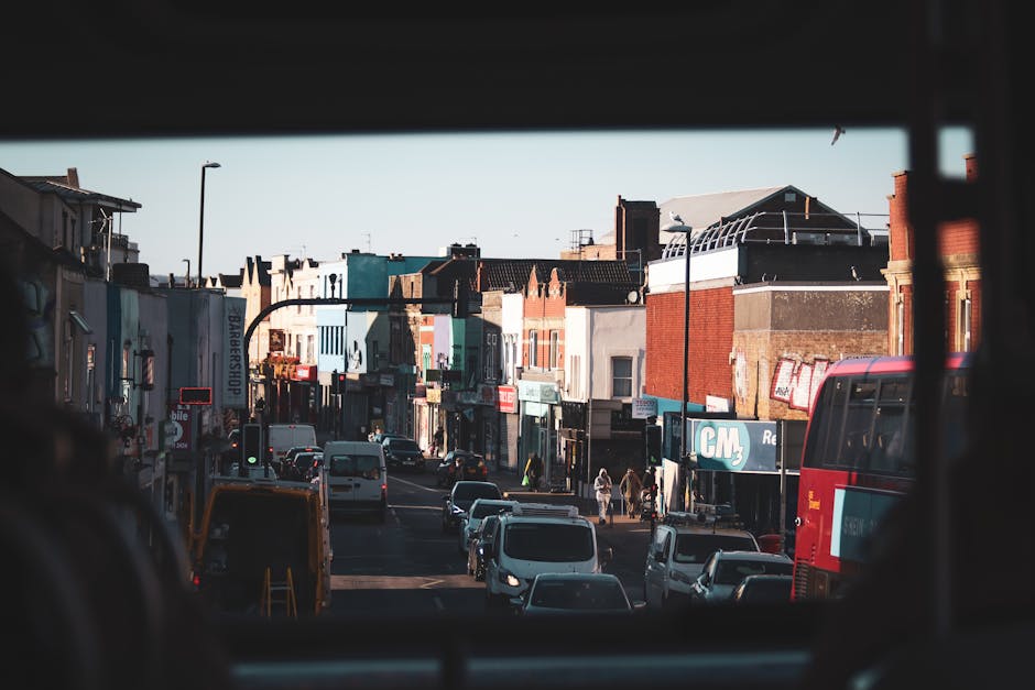 View of a busy Woolwich street captured from inside a vehicle, with the window framing the scene. The street is lined with a variety of small shops and commercial buildings, some with colorful signage and storefronts. Several vehicles, including white vans, a red double-decker bus, and passenger cars, are either parked along the curb or moving through the street. A streetlamp and utility poles are visible above the buildings, and pedestrians are walking along the sidewalk. The scene indicates active urban traffic and foot traffic typical of a home relocation and furniture transport environment, where loading and unloading might occur near the curb, supporting local removal and moving services such as those offered by Man With a Van Woolwich. The bright daylight enhances visibility of the street scene, highlighting the typical setting for a house removals operation involving careful packing, loading, and transport of household items.