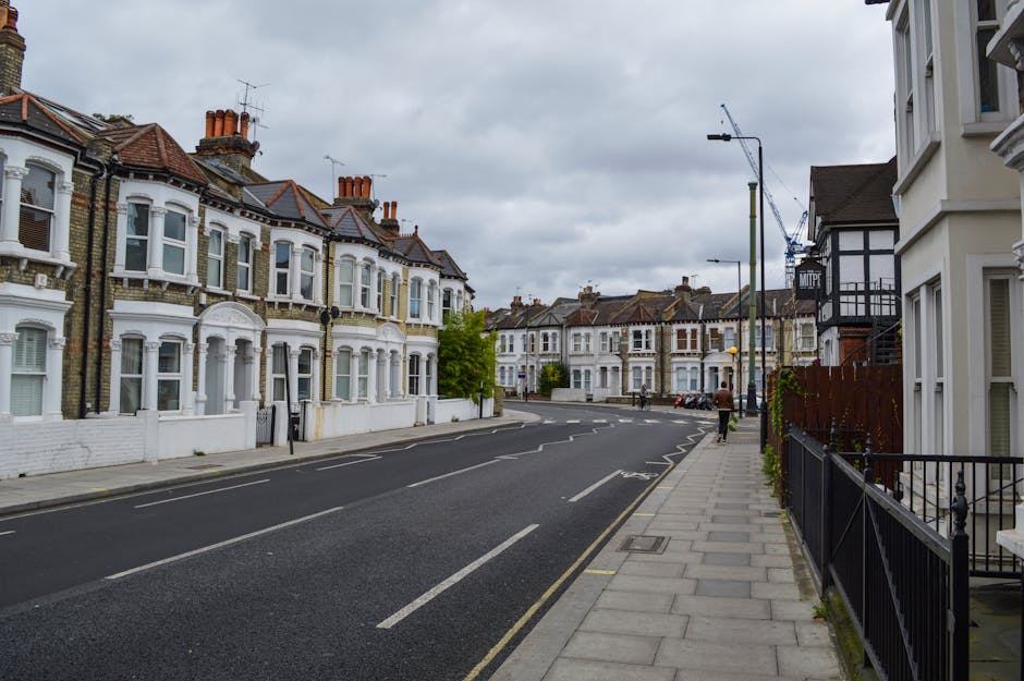 A quiet residential street in Woolwich with a row of Victorian-style terraced houses on the left, featuring white-painted facades, large bay windows, and red-tiled roofs. The street has a paved sidewalk on the right side, separated from the road by a black metal railing, with a few small trees planted along it. The road is mostly clear of traffic, with a few parked cars visible in the distance, and a person walking along the sidewalk wearing a brown jacket. The sky appears overcast, with grey clouds covering the scene. This setting may relate to home relocation or furniture transport operations, as part of a house removals process, with the environment suitable for loading or unloading activities near dwellings.