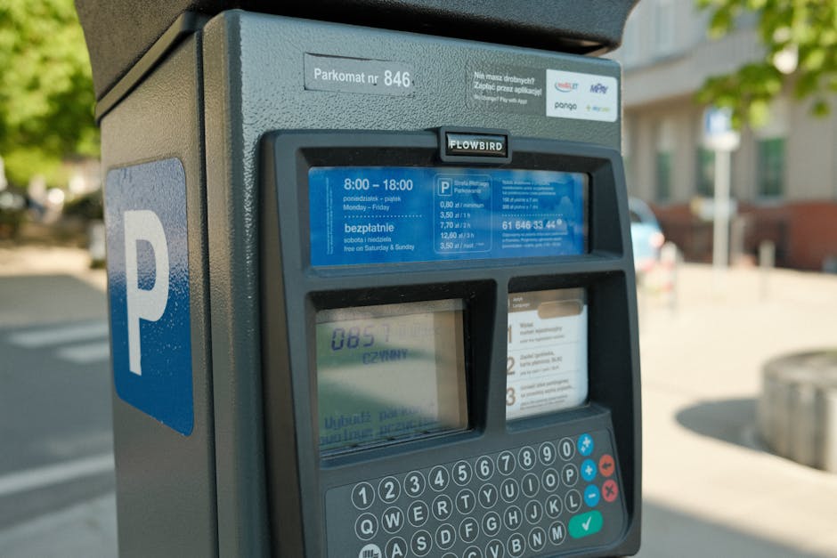 Close-up image of a parking meter situated on a pavement in Woolwich, featuring a predominantly grey body with a large blue 'P' symbol on the side indicating parking. The digital display on the meter shows parking payment details, including a start time of 8:00 and an end time of 18:00 from Monday to Friday, with information about free parking on weekends. The front panel includes operating instructions, a keypad with numbers and function keys, and a small screen displaying transaction information. In the background, blurred residential buildings and greenery suggest an urban neighbourhood environment. This parking meter, used for managing parking permits during home relocation or furniture transport, is associated with Woolwich Council parking regulations. Occasionally, professional removal teams, like those from Man With a Van Woolwich, utilize such parking options when carrying out house removals and moving services within the area, ensuring compliance with local parking policies during loading and unloading processes.