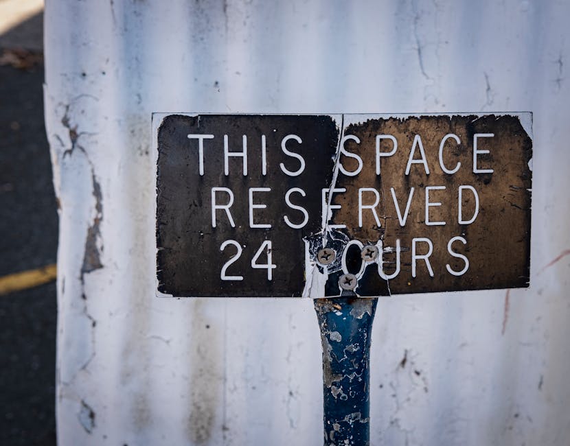 A close-up of a weathered, rectangular metal parking restriction sign mounted on a blue pole, attached with rusted screws. The sign displays the message 'THIS SPACE RESERVED 24 HOURS' in white capital letters on a black and brown background, with visible peeling paint and corrosion indicating exposure to the elements. Behind the sign, there is a white, textured wall with cracks and chipped paint, and a dark asphalt surface with a faint yellow line is partially visible on the left side. The environment suggests an urban setting, relevant for house removals and moving logistics, as used by Man With a Van Woolwich for residential furniture transport and home relocation services.