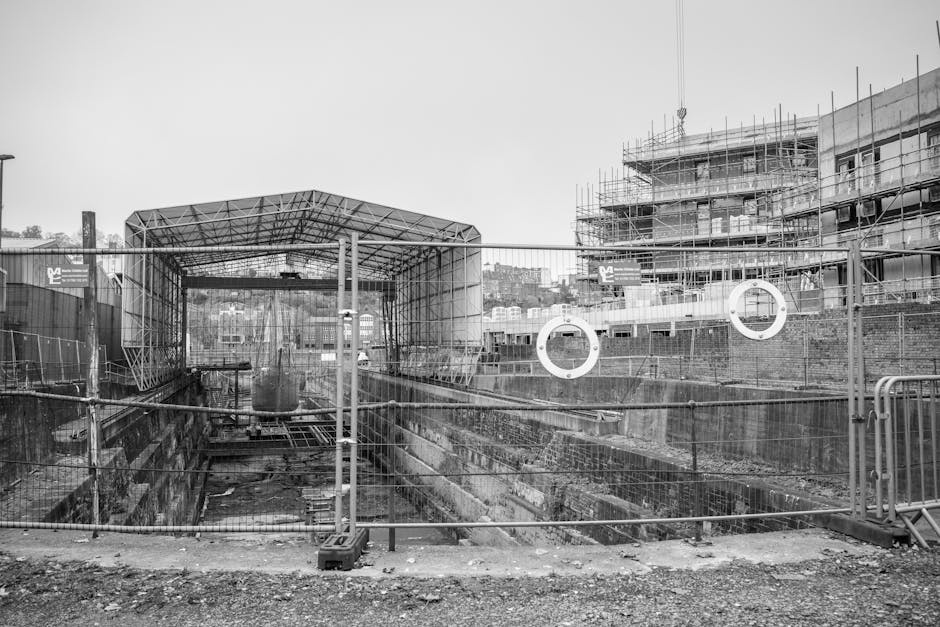 A black-and-white photograph of an urban construction site featuring a depth of field that shows a partially excavated area with reinforced concrete and brick walls, surrounded by safety fencing with metal posts and wire mesh. Inside the site, there is a large, temporary metal structure with a curved roof, possibly a shelter or work platform, positioned at the entrance. The ground within the construction zone consists of dirt and debris, with construction materials scattered around. To the right, a multi-storey building under construction is visible, covered in scaffolding and partially finished with concrete floors and exterior walls. In the background, residential buildings and a hilly landscape are visible, indicating the site is within an established urban area. The scene appears to be during daylight hours and is part of a home or building renovation or new build project located near a body of water, consistent with the location details of Woolwich Dockyard, as referenced on the webpage for Man With a Van Woolwich’s removals service.