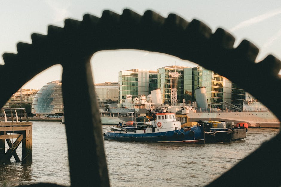 A view of a river with several boats of varying sizes and colors, including a small blue and white tugboat in the foreground and larger ships docked alongside modern glass-fronted office buildings in the background, framed by a large, dark, toothed gear-shaped structure in the foreground. The scene captures a shipping and transportation setting possibly related to home relocation or furniture transport, with the daylight illuminating the water and surrounding structures. The boats are moored near a quay with wooden posts and metal railings on the left side of the image, highlighting active maritime operations that are part of a house removals or loading process. The modern, reflective buildings suggest a city waterfront environment, where equipment such as trolleys or moving vans operated by companies like Man With a Van Woolwich might facilitate furniture and packing material movement, especially in a narrow access scenario typical of an urban move, as indicated by the PAGE_TITLE.