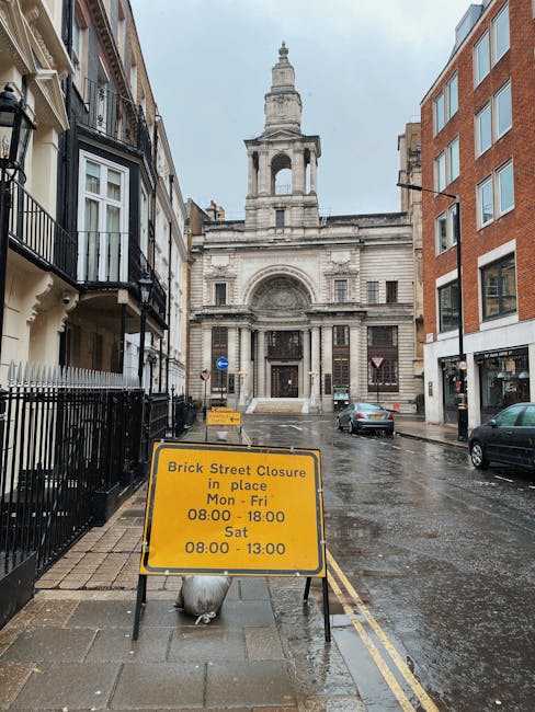 A wet, narrow street in Woolwich during daytime with rain-slicked pavement and parked cars on both sides. On the left, there are residential buildings with black wrought-iron balconies and cream-colored facades. The right side features modern red-brick buildings with large windows. At the end of the street, a historic stone church or government building with a tall central tower and classical architectural details is visible. In the foreground, a yellow road closure sign stating 'Brick Street Closure in place Mon - Fri 08:00 - 18:00, Sat 08:00 - 13:00' is positioned on the sidewalk, secured by a white bag at its base. This scene illustrates an urban environment where house removals or furniture transport activities could be temporarily affected by road closures, and the image is associated with professional removals services like those offered by Man With a Van Woolwich.