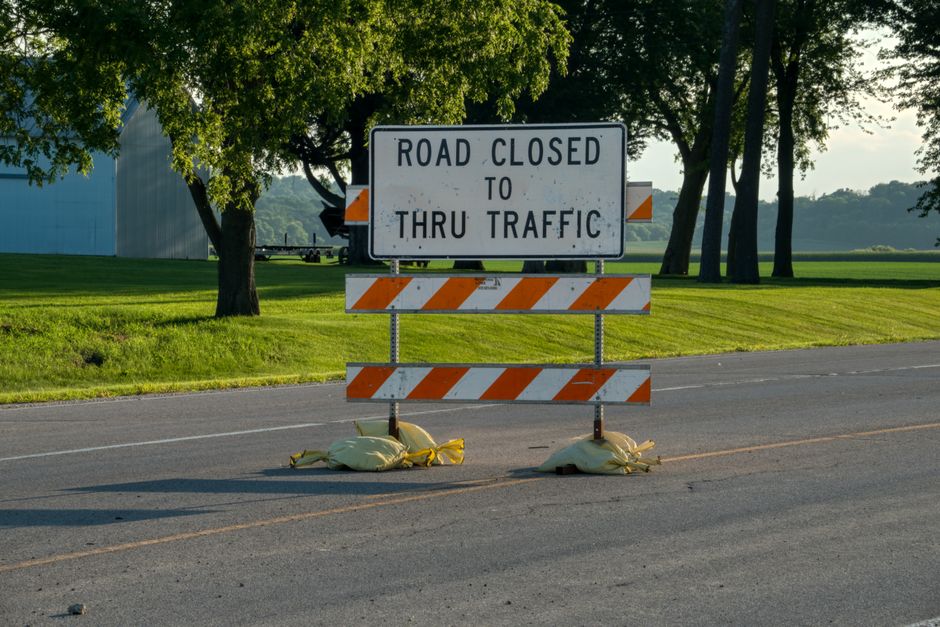 A road closure sign is positioned in the middle of a paved street, supported by two sandbag weights at its base. The sign reads 'ROAD CLOSED TO THRU TRAFFIC' in black lettering on a white background, with orange and white striped barricades below, indicating temporary road restrictions. In the background, there are green trees with dense foliage, casting shadows on the grass and pavement, suggesting a daytime outdoor setting. This scene may be associated with road closure management during a home relocation or moving process, as part of logistical planning by a professional removals service like Man With a Van Woolwich. The open space and natural environment emphasize the importance of traffic coordination in residential moving projects, where access and transportation routes are carefully managed.